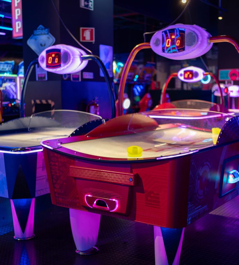 Modern air hockey tables inside a neon-lit shopping mall, complemented by various gaming machines in an illuminated arcade. Entertainment concept.