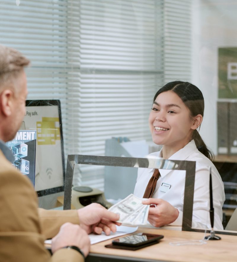 Caucasian middle aged man handing cash to young adult bank teller behind counter, both interacting at branch, woman smiling while processing transaction, glass partition visible