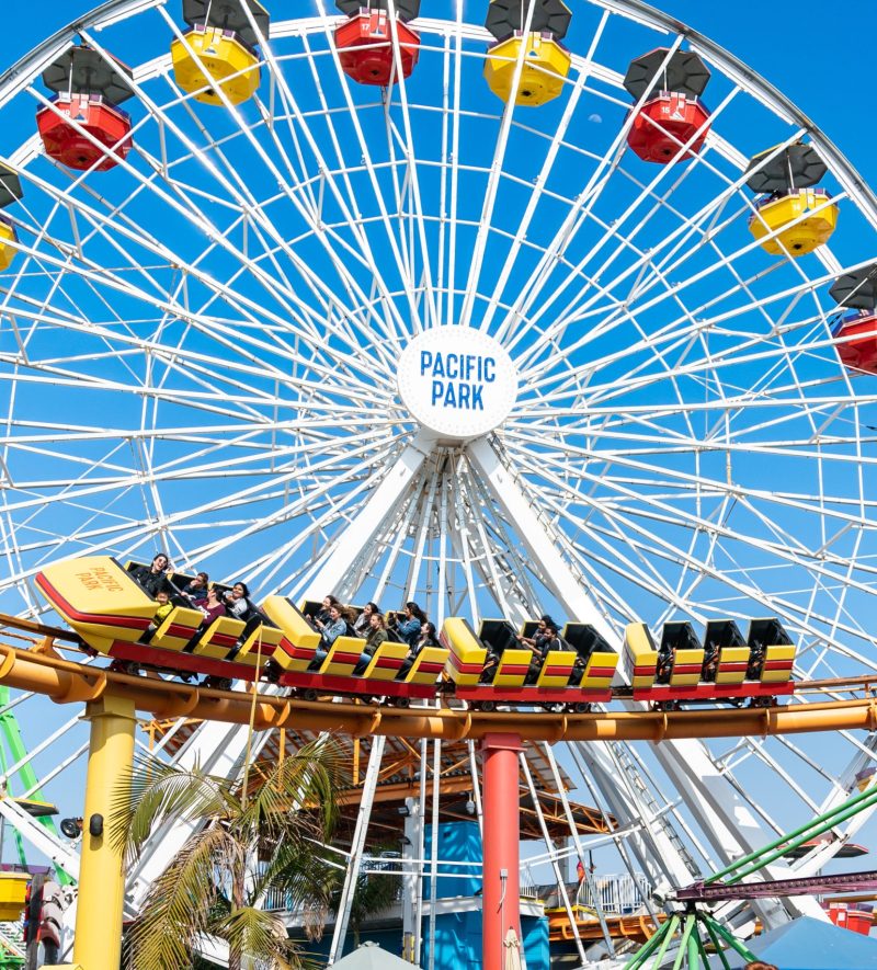 California, USA - May 22, 2018: Ferris Wheel with pacific park logo at center and people riding roller coaster in amusement park at santa monica pier on sunny day