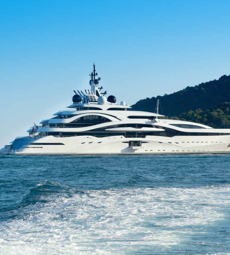 Huge luxury motor yacht cruising offshore in a calm ocean passing a forested coastline viewed over the wake of a passing ship