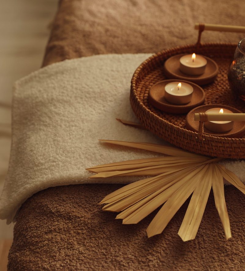 Wicker basket with burning candles, dry palm leaf and fresh towels on massage table in spa center, closeup